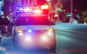 A police SUV with a full light bar across its roof blazing in alternating red and blue emergency lights sits head-on in the middle of a rain-slicked urban street at night its bright white headlights and additional blue grille lights illuminating the wet pavement in front of it with a colorful blur of city storefronts- neon signage- traffic signals- and streetlights glowing in the bokeh background on both sides of the road — the commanding presence of a law enforcement vehicle conducting a DUI traffic stop on a busy city street after dark in the Upstate of South Carolina.