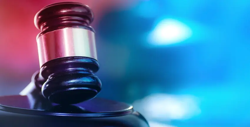 A dark wooden judge's gavel with a wide silver metallic band sits upright on its matching dark circular sound block photographed at close range from a low angle against a dramatically lit background awash in vivid red light on the left side and bright electric blue light on the right — the contrasting red and blue tones unmistakably evoking police emergency lights directly connecting the authority of the courtroom to the legal consequences a driver faces after being charged during a DUI traffic stop and could use help from DUI Lawyer Greenville.
