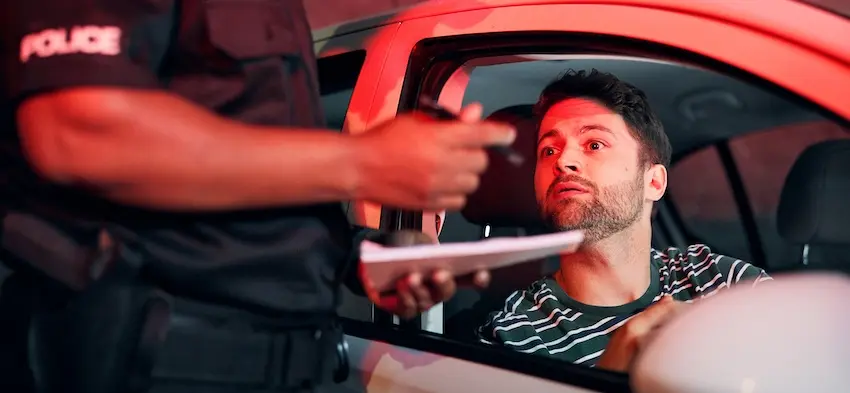 A male driver with dark curly hair and a short beard wearing a green and white striped shirt sits in the driver's seat of a red vehicle with the window rolled down- his expression wide-eyed and tense as he looks up at a police officer standing outside the car door- the officer's black uniform with "POLICE" printed across the chest visible from the torso down as he extends one hand toward the driver and holds a clipboard or citation pad in the other the entire scene bathed in deep red emergency light — capturing the high-pressure interaction between a motorist and law enforcement during a DUI traffic stop in the upstate of South Carolina.
